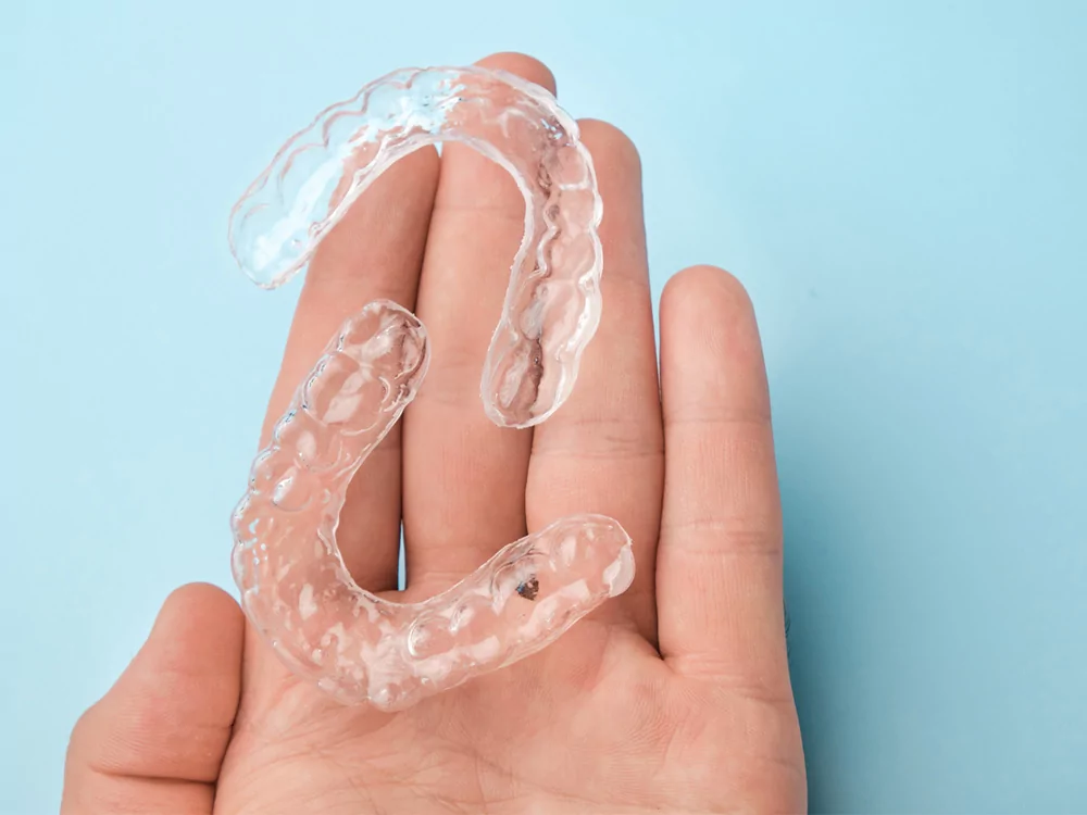 The image shows a palm with a pair of clear dental retainers on it. The background color is baby blue.