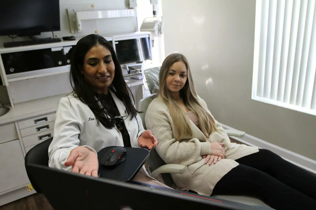 An image depicting two individuals in a healthcare setting, likely a dental office. One, a professional in a white coat, gestures while interacting with a seated patient. A computer monitor and medical equipment are visible in the background.