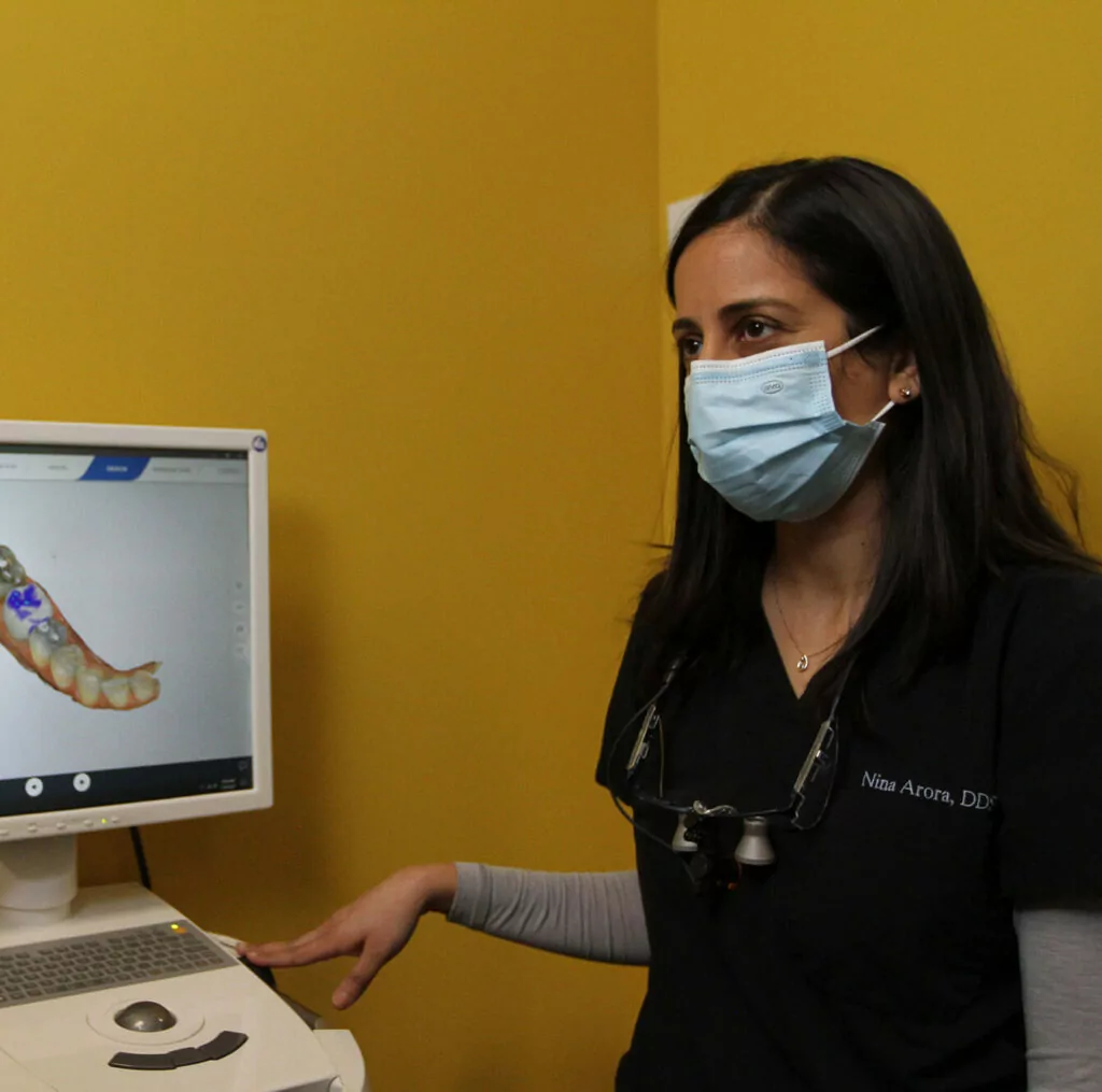 A dental professional in a black shirt with "Nina Arora, DDS" embroidered on it stands next to a monitor displaying a 3D rendering of a dental model. They wear a face mask and dental loupe, with a yellow wall in the background.