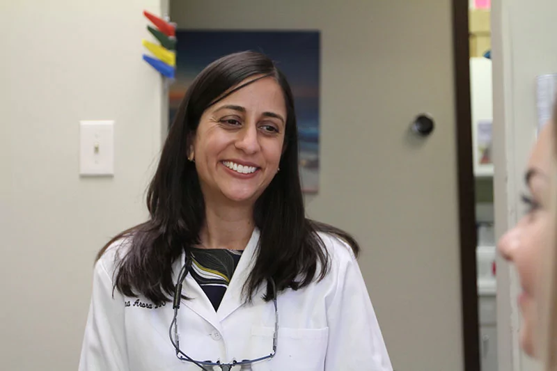 Dr. Nina Arora of Romeoville Dental Center n a white lab coat with embroidered text, wearing glasses, stands in a professional or medical setting. The background features a wall with a light switch, colorful object, painting, and doorway.