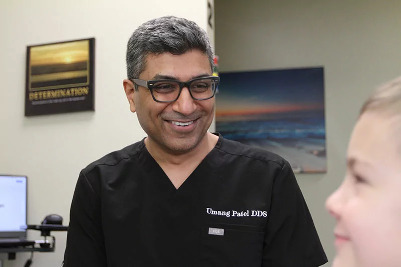 The image features a person wearing a black scrub top with "Umang Patel DDS" embroidered on it, indicating a professional setting, likely a dental office.