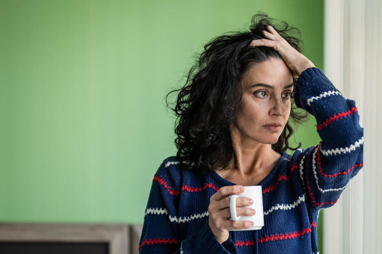 A person with dark curly hair wearing a blue striped sweater sits indoors against a light green wall, holding a white mug and resting one hand on their head, appearing thoughtful or concerned—suggestive of someone reflecting on dental emergencies such as sudden tooth pain, cracked teeth, or urgent dental care needs.